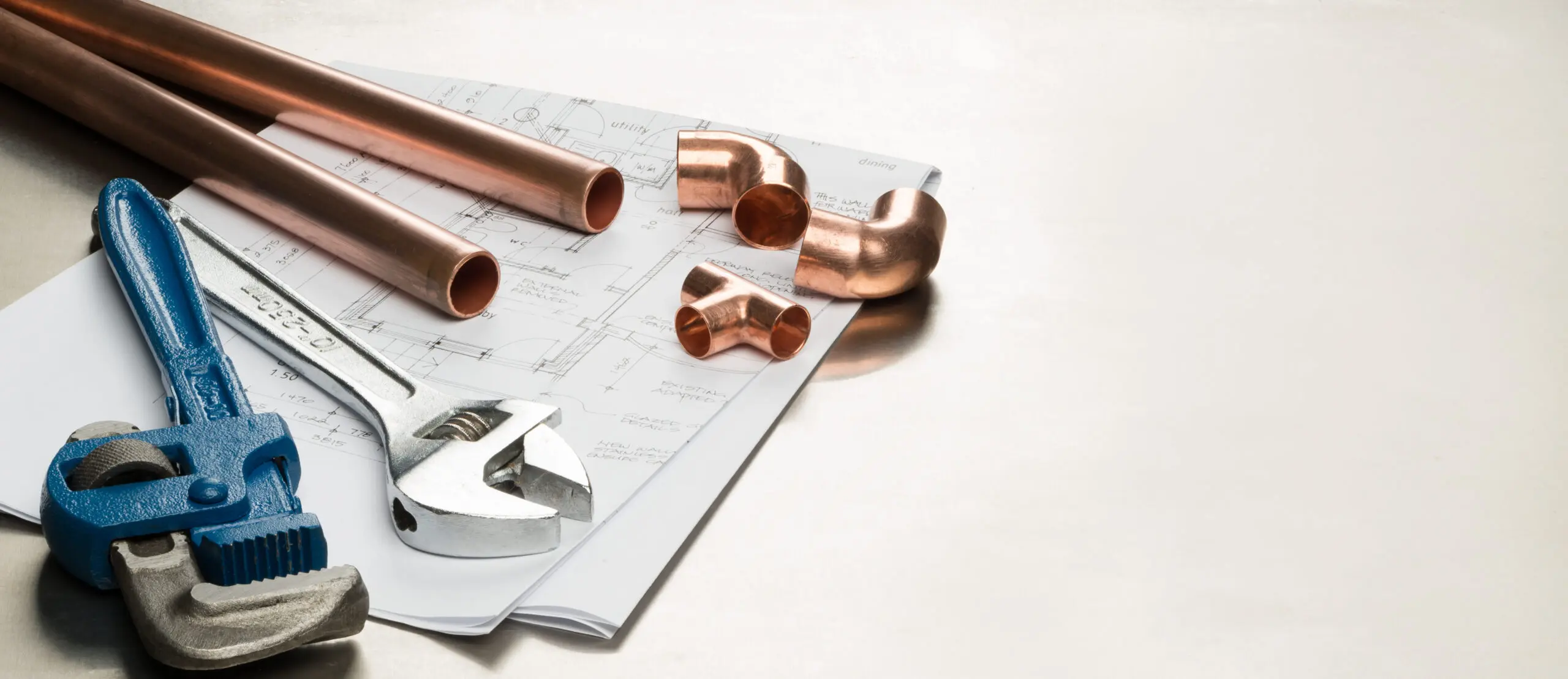 A blue pipe wrench, adjustable wrench, copper pipes, and fittings lie on architectural blueprints on a light surface, ready for the skilled hands of a plumber.
