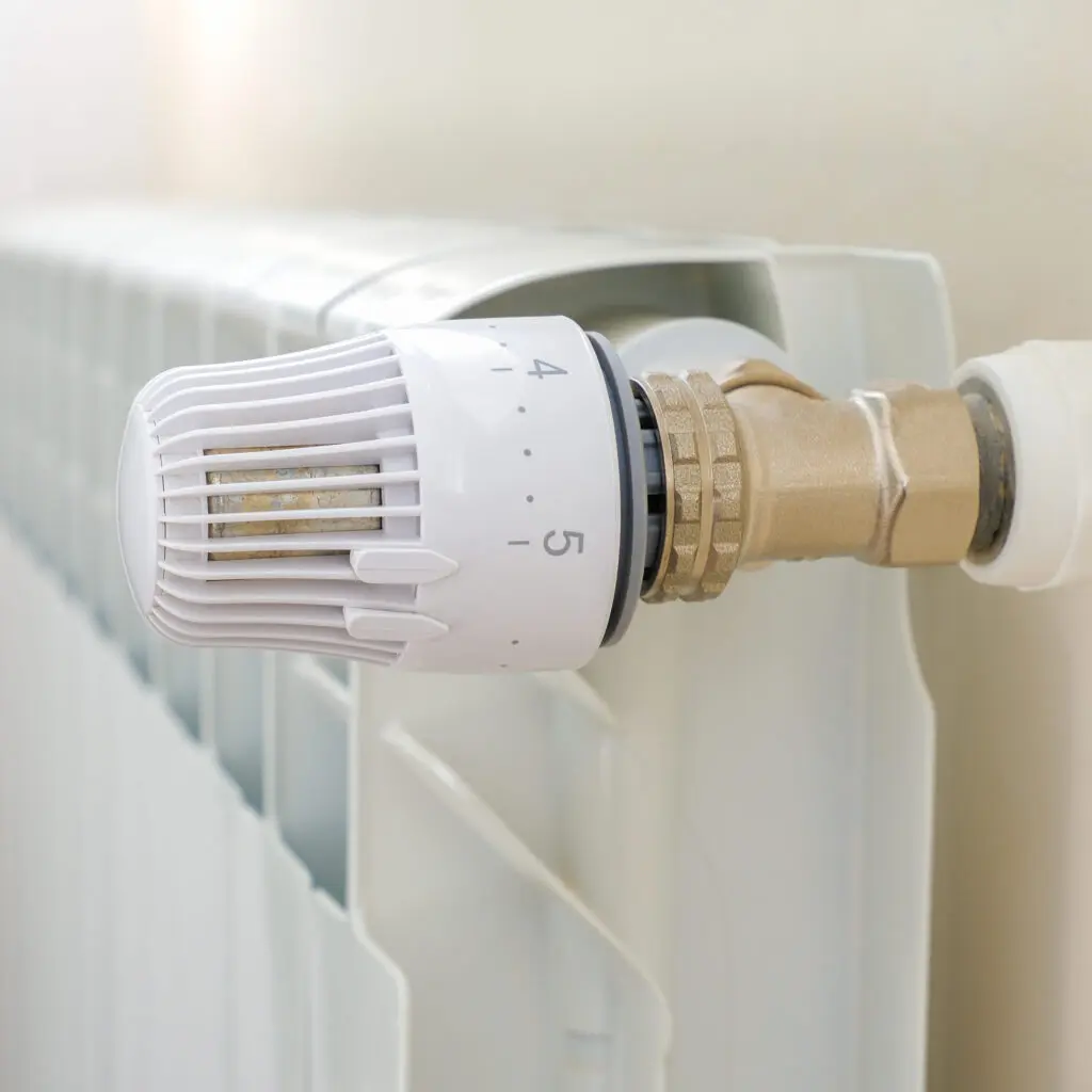 A close-up of a white radiator with a numbered temperature control valve, set between 4 and 5, attached to a metal pipe. The background is softly blurred.