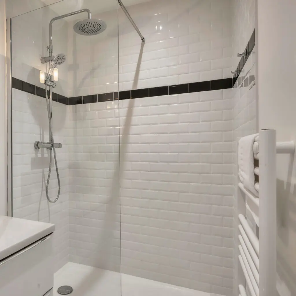 Modern bathroom shower with white subway tile walls, a black accent tile stripe, a glass partition, a rainfall showerhead, and a towel warmer with folded white towels.