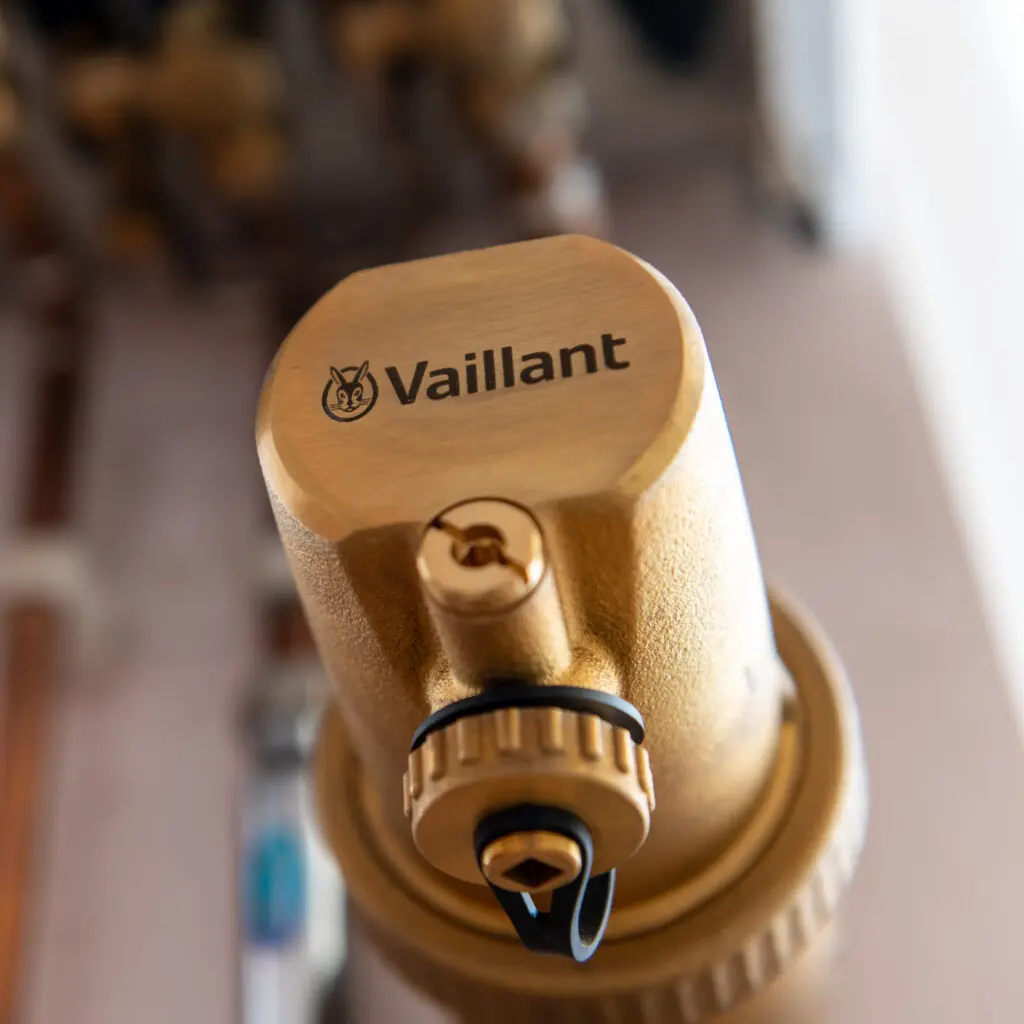 Close-up of a brass mechanical component with the brand "Vaillant" and a logo engraved on top, featuring a screw and a black plastic loop at the base, likely part of a heating or plumbing system.