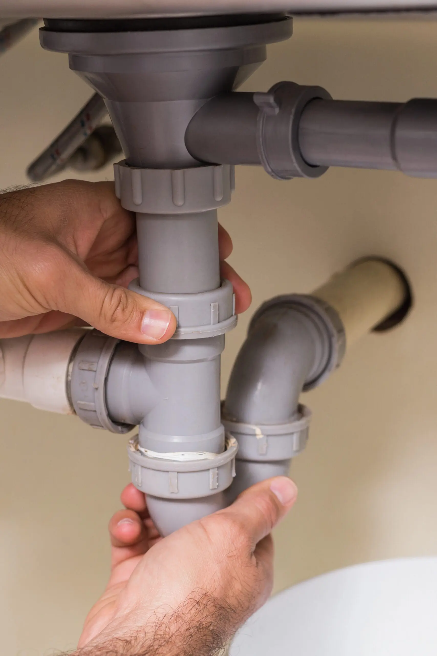 A person’s hands are assembling or fixing a gray plastic pipe under a sink, connecting multiple sections of plumbing.